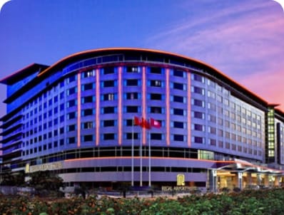 The Regal Airport Hotel features a modern design with illuminated curves, set against a twilight sky, showcasing flags out front.