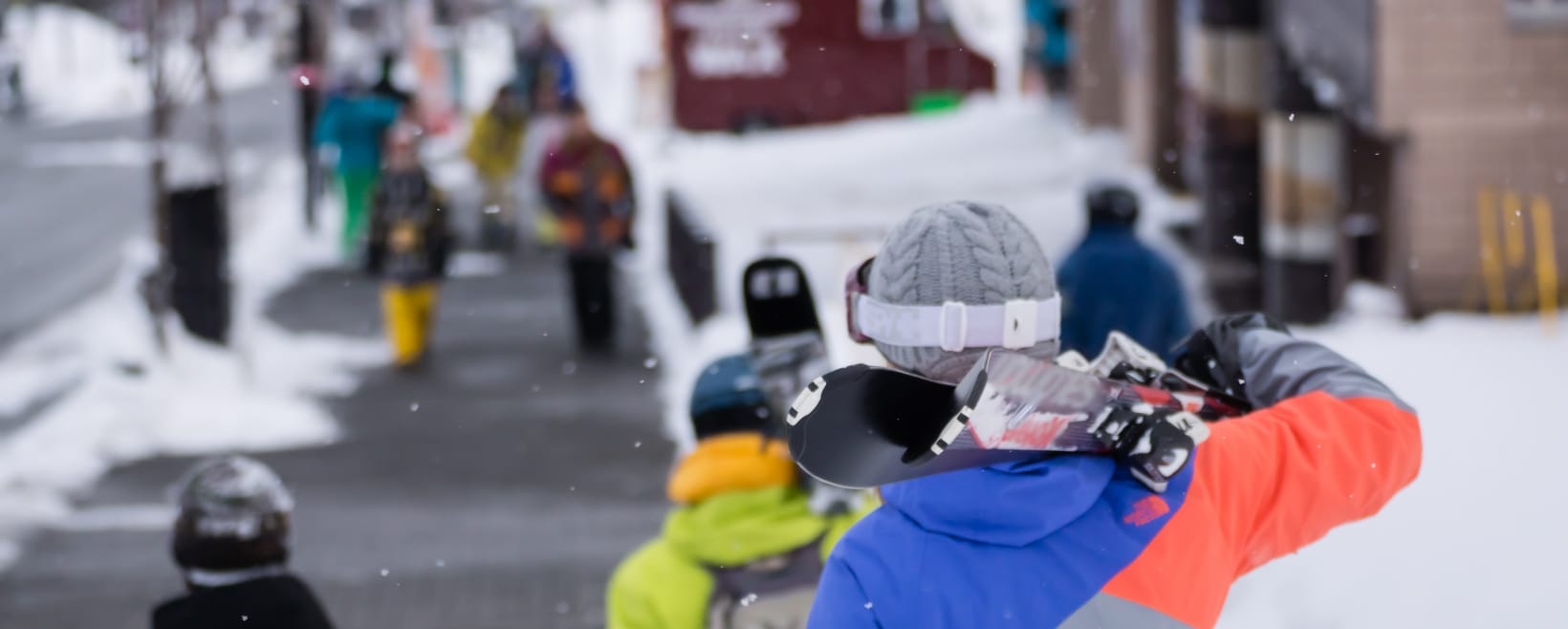 A group of people walking in the snow, one person carries a snowboard, wearing colorful winter attire against a snowy backdrop.