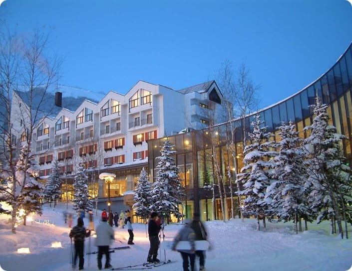 People walking near snow-covered trees in front of a lit building at dusk.